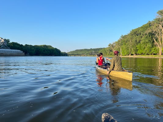 a pair of canoeists paddle down a river with industrial infrastructure on the left bank, leafy forest on the right and a rail bridge in the far distance