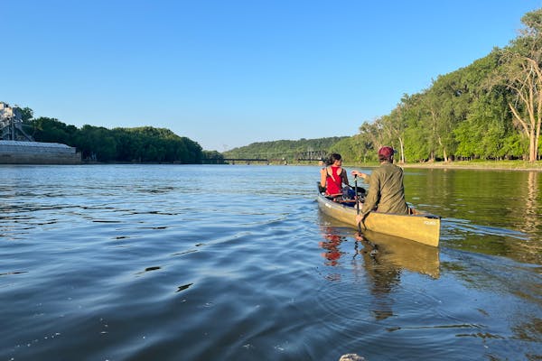 a pair of canoeists paddle down a river with industrial infrastructure on the left bank, leafy forest on the right and a rail bridge in the far distance