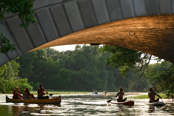 paddlers on canoes and paddleboards make their way under the arch of a concrete bridge with lush green leafy trees in the background