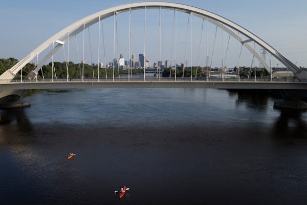 two kayakers are seen from above as they pass under the large white arches of a bridge with the skyline of downtown Minneapolis visible in the distance