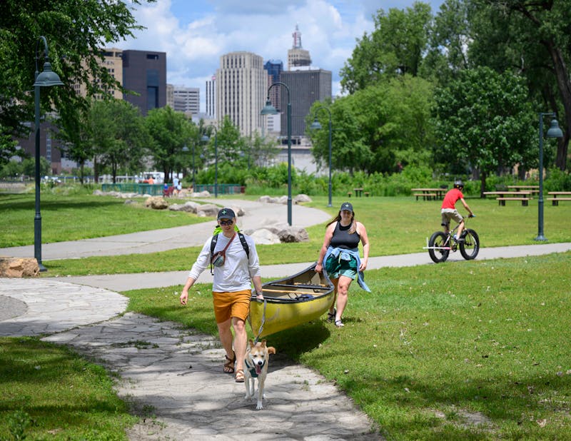 Two adults carry a yellow canoe on land, with a yellow dog in front. 